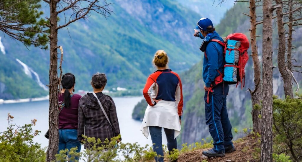 Hiking guide pointing towards the village of Mo from a scenic viewpoint, with three hikers enjoying breathtaking fjord and mountain views.