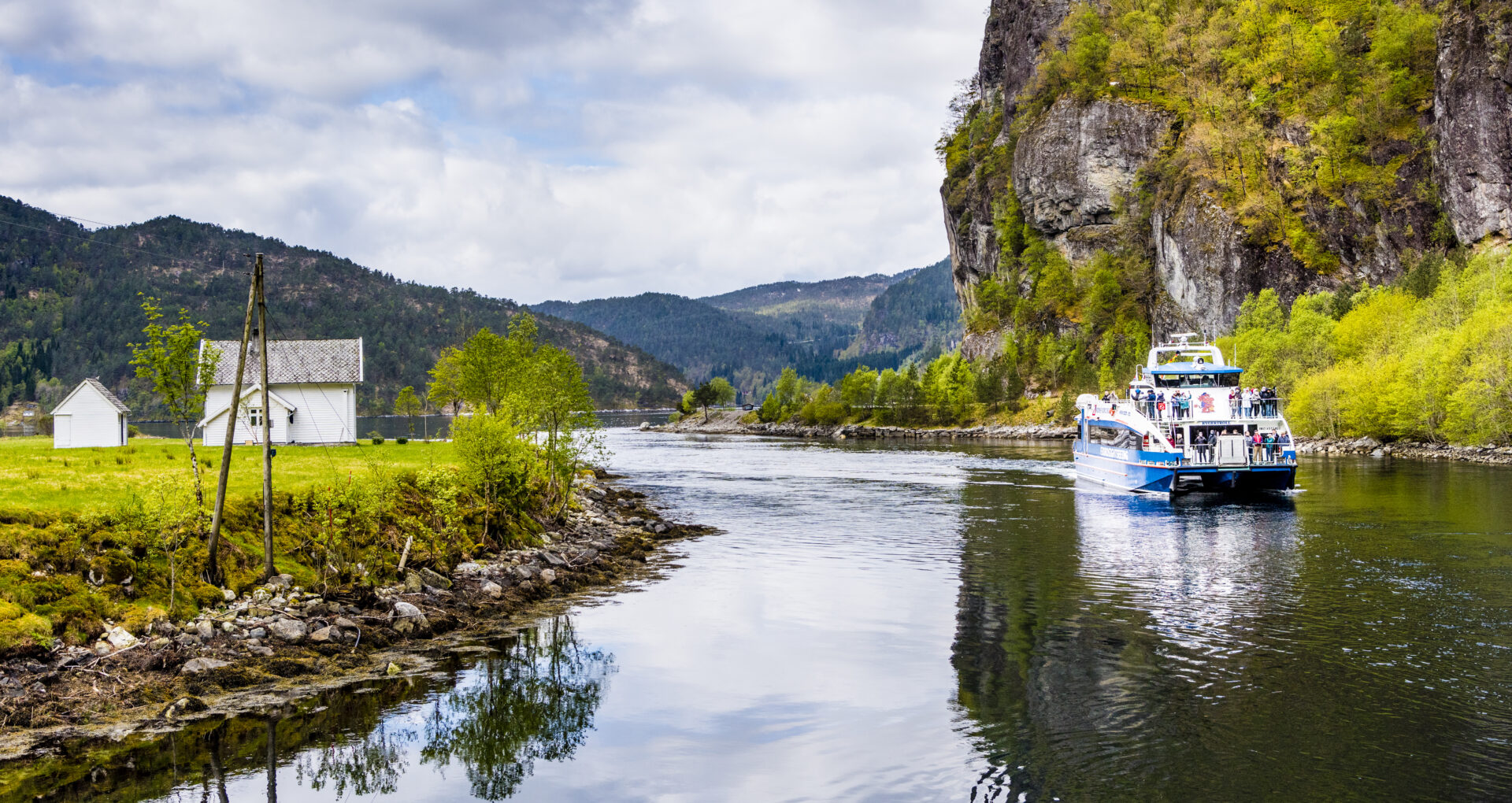 A modern fjord cruise catamaran gliding through Mostraumen straits with green hills and forests in the background near Bergen, Norway.