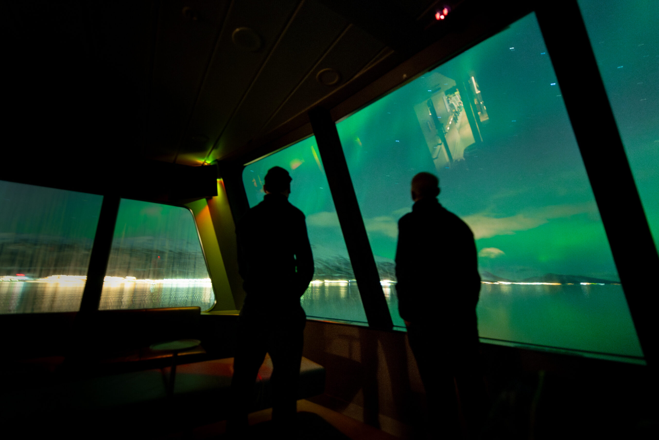 To menn som nyter synet av nordlyset over Tromsøysundet. Store panoramavinduer i oppvarmet salong. Mennene er med på en nordlystur med båt fra Tromsø.