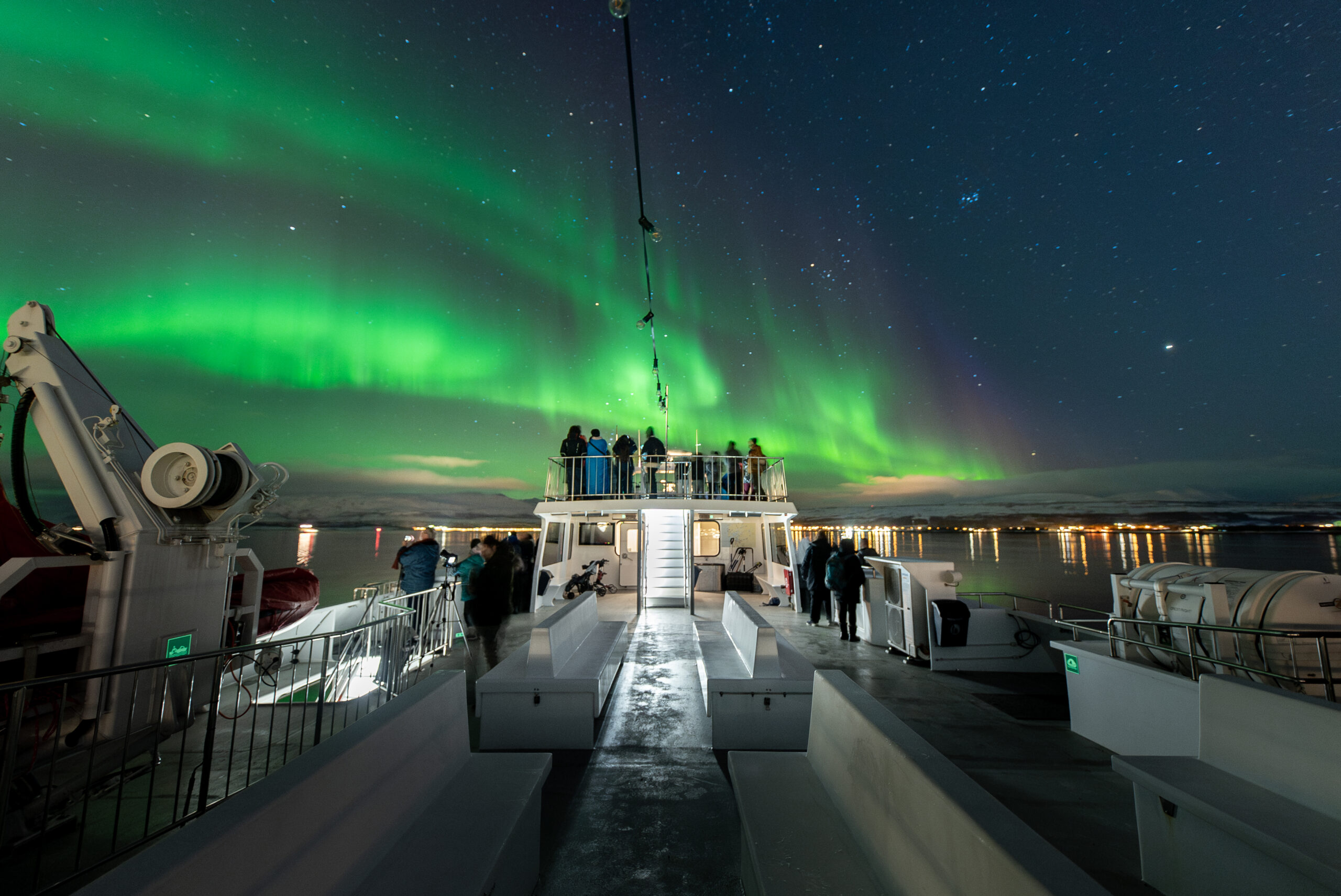 En gruppe mennesker har trukket ut på dekket under Nordlys båttur fra Tromsø. Stjerneklar himmel og dansende grønt nordlys med Tromsø by i bakgrunnen