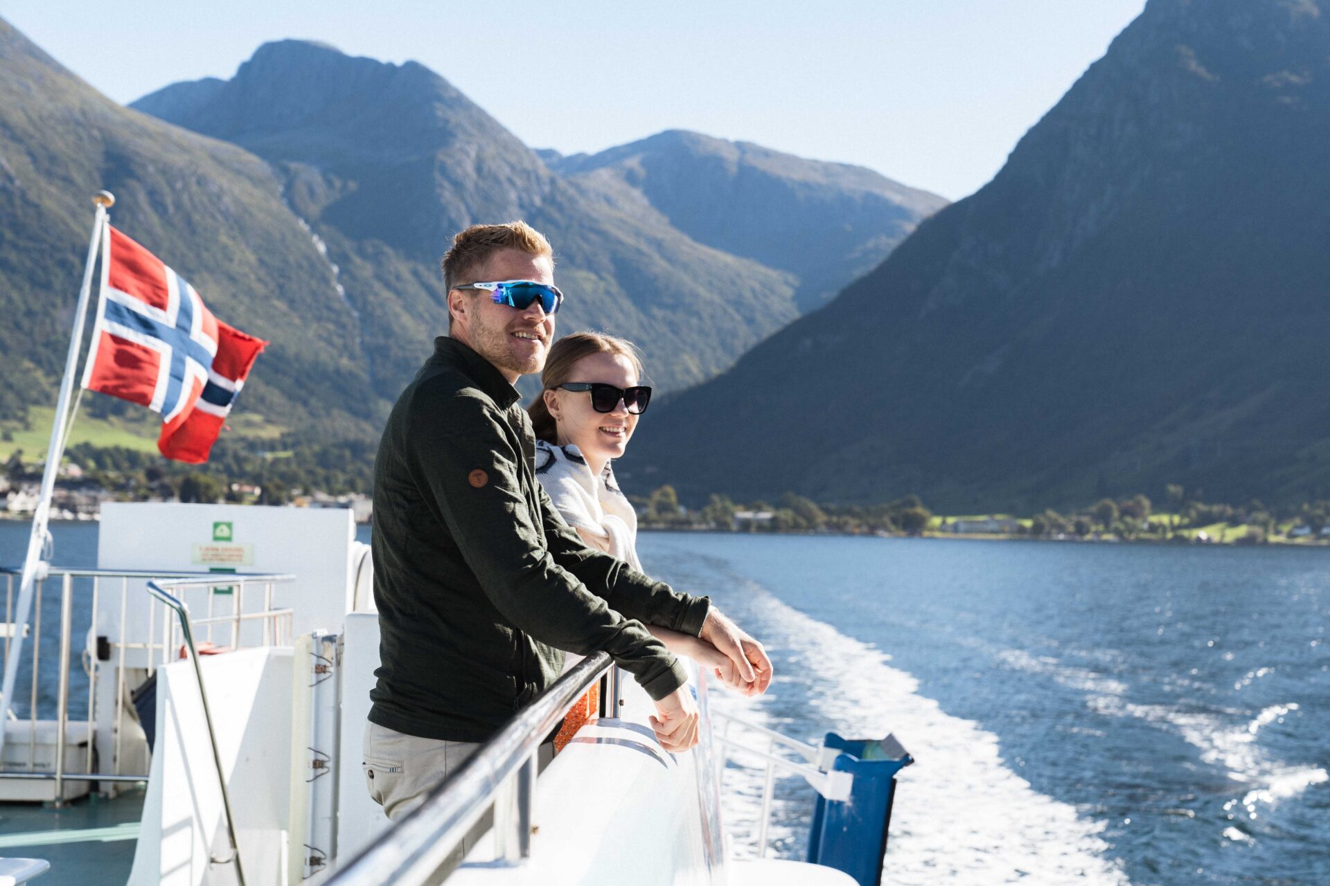 A couple standing on the outdoor viewing deck of the Hardangerfjord Express, enjoying the stunning fjord scenery as the boat departs from Rosendal towards Bergen