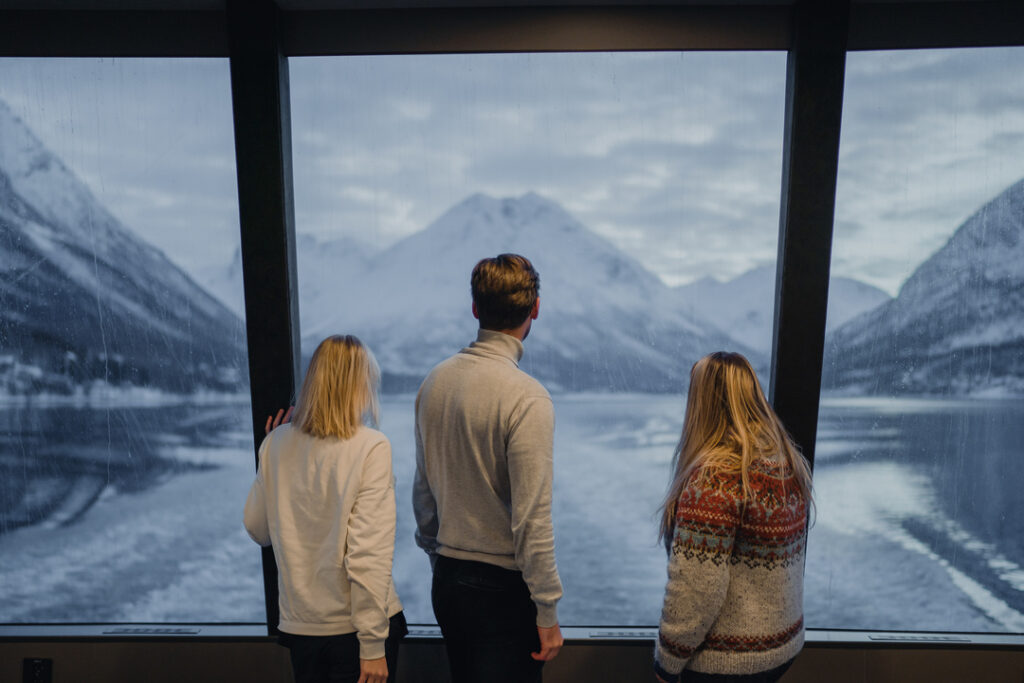 Tre gjester nyter panoramautsikten fra bakre del av Rødnesbåten under fjordcruise i Tromsø, med Lyngenalpene i bakgrunnen.