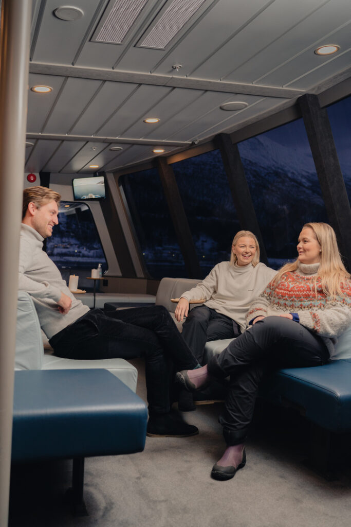 Tre gjester sitter komfortabelt bakerst i Rødnesbåten med panoramavinduer under fjordcruise i Tromsø, omgitt av smil og latter.
