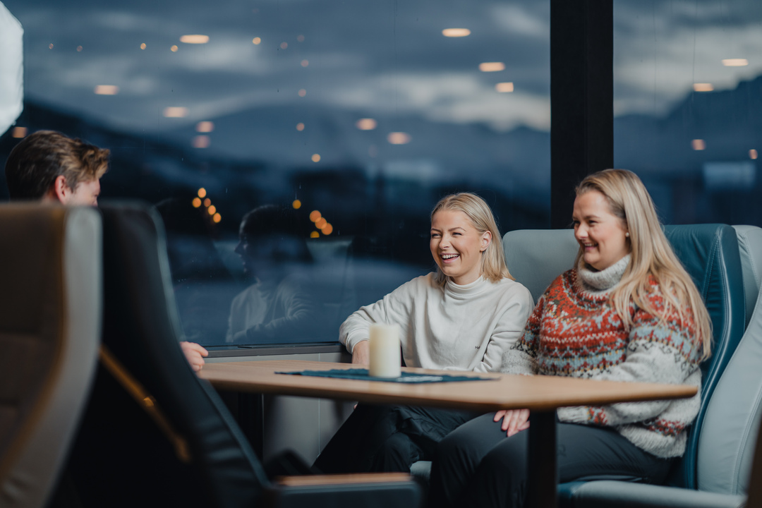 Tre gjester sitter komfortabelt i den oppvarmede salongen på Rødnes fjordcruise i Tromsø, med panoramautsikt over fjorden gjennom store vinduer.