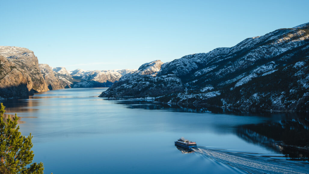 Rygerelektra, elektrisk båt seiler stille gjennom den majestetiske Lysefjorden i vinterlys, omgitt av snødekte fjell og speilblankt vann. Opplev fjordens magi på et bærekraftig fjordcruise fra Stavanger.