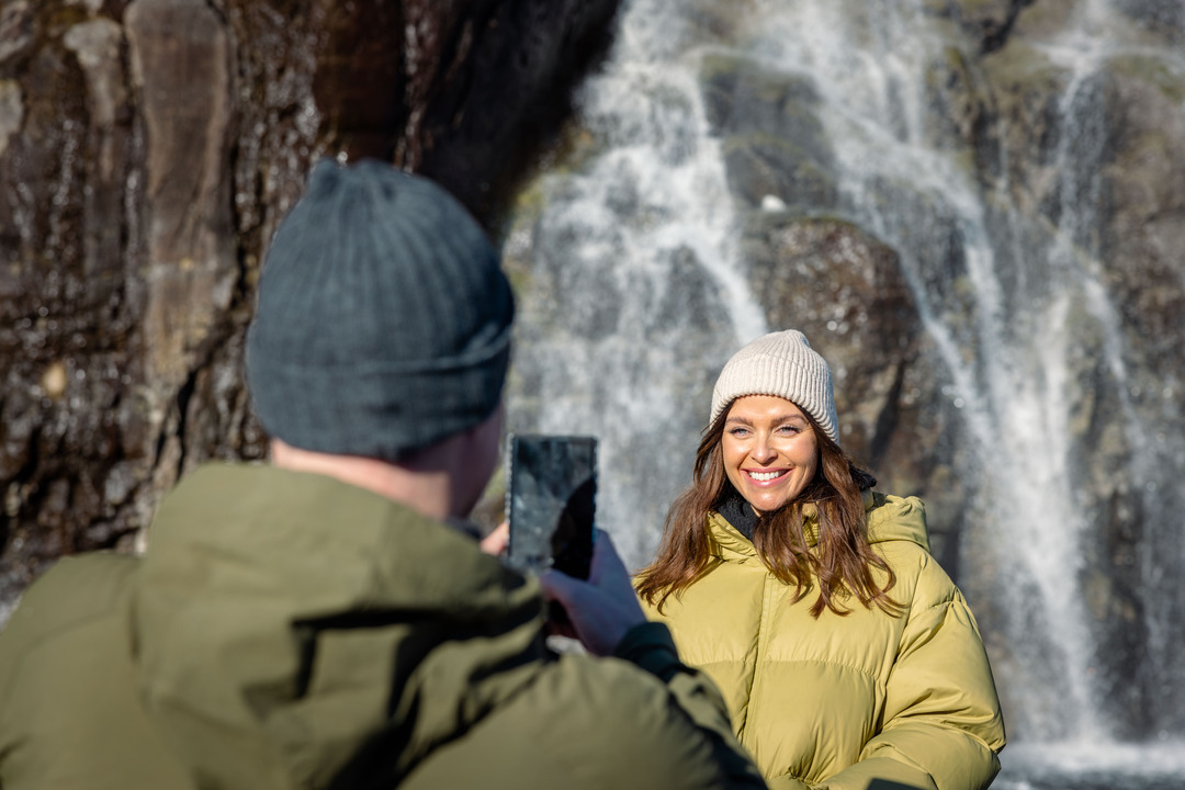 En kvinne i varm jakke smiler mens hun står nær en brusende foss på Lysefjorden, mens en mann tar bilde.