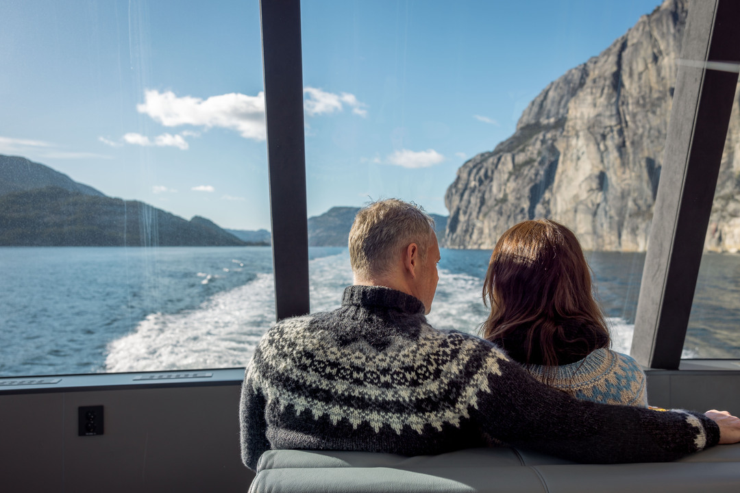Et par i norske ullgensere nyter utsikten over de høye fjellveggene i Lysefjorden gjennom store panoramavinduer.