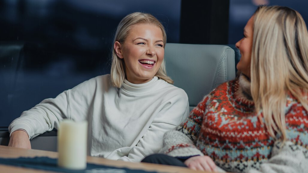 Two smiling women dressed in warm sweaters are enjoying a cozy moment indoors on a Northern Lights cruise from Tromsø. A candle adds to the warm and relaxing atmosphere as they share laughter, with the Arctic night visible through the window.