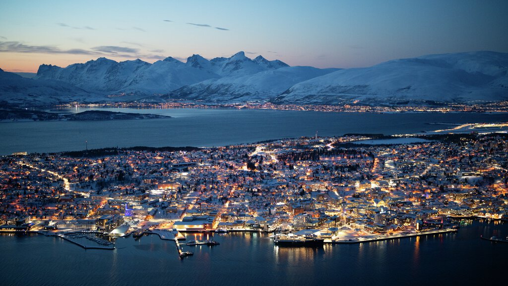 A breathtaking aerial view of Tromsø at dusk, with the city's warm lights glowing against the snowy Arctic landscape. The fjord reflects the last light of the day, while majestic mountains stand in the background, creating a stunning winter scene.