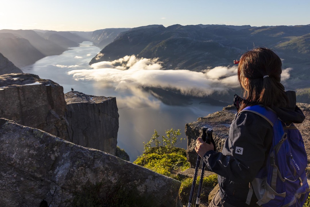 A hiker overlooking the Preikestolen Platau and Lysefjorden on a early morning with the sun shining trough and low clouds hanging over Lysefjord.