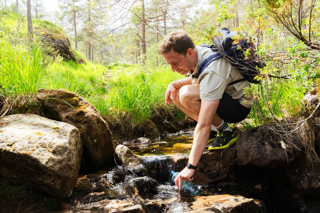 Hiker refilling a water bottle from a fresh mountain stream on the way to Slottet, dressed in proper hiking gear. Practicing Leave No Trace principles in the Norwegian fjords.