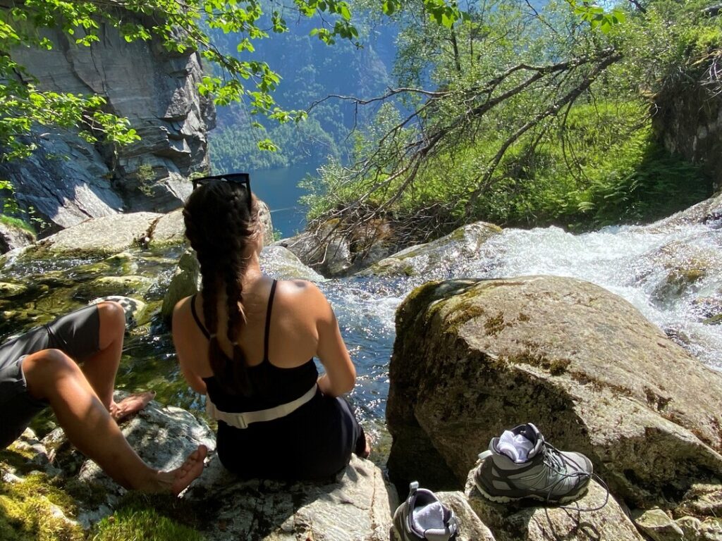 Two hikers cool their feet in a mountain stream during a hike in Bergen, enjoying a breathtaking fjord view on a sunny summer day.