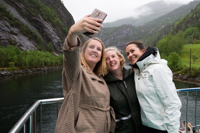 Tre kvinner smiler og tar en selfie på dekk under et fjordcruise fra Bergen til Mostraumen.