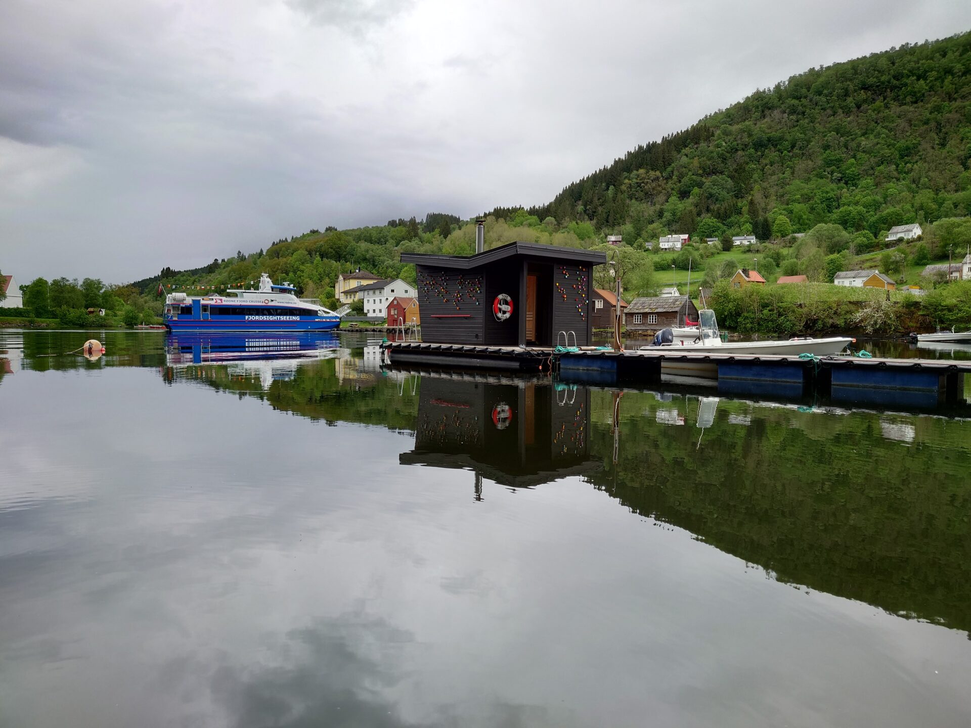 Floating sauna on a calm fjord surrounded by lush green hills in Hosanger
