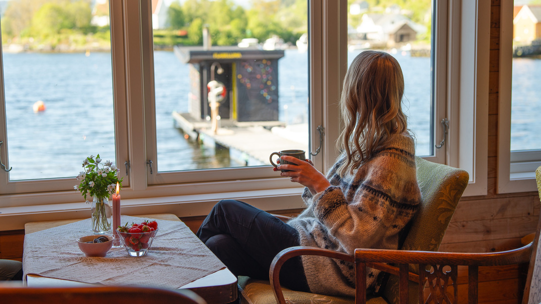 Dame som nyter en varm kopp kaffe hos Den Grøne Fabrikken, sitter og nyter utsikten over Mjøsvågen, dampbastuen og Osterfjorden