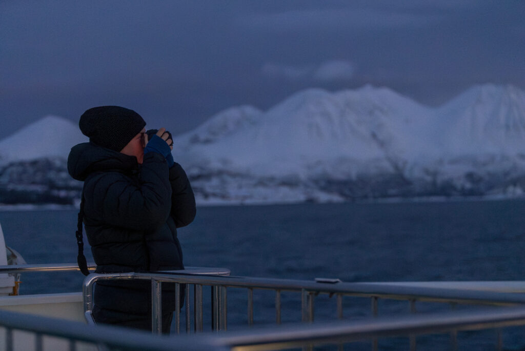 A guest taking a photo of the Arctic fjords during early dusk in the polar night, with calm waters and snow-covered mountains near Tromsø in the background.