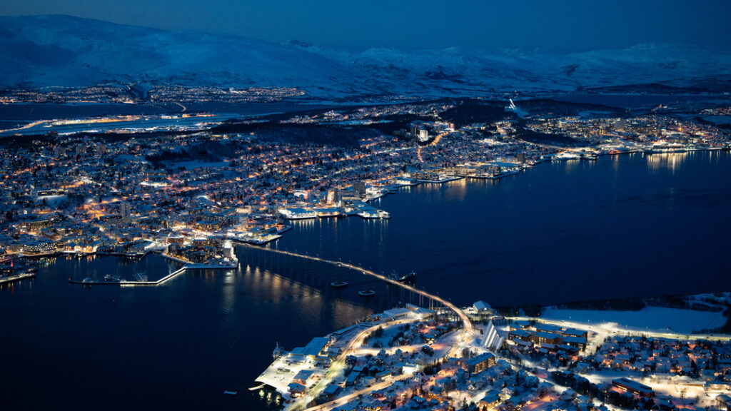 Panoramic winter view of Tromsø city and Arctic fjords at dusk from Fjellheisen viewpoint, with glowing city lights, the airport in the distance, and fjords surrounding the city center. Rødne fjord cruises operate in these waters.
