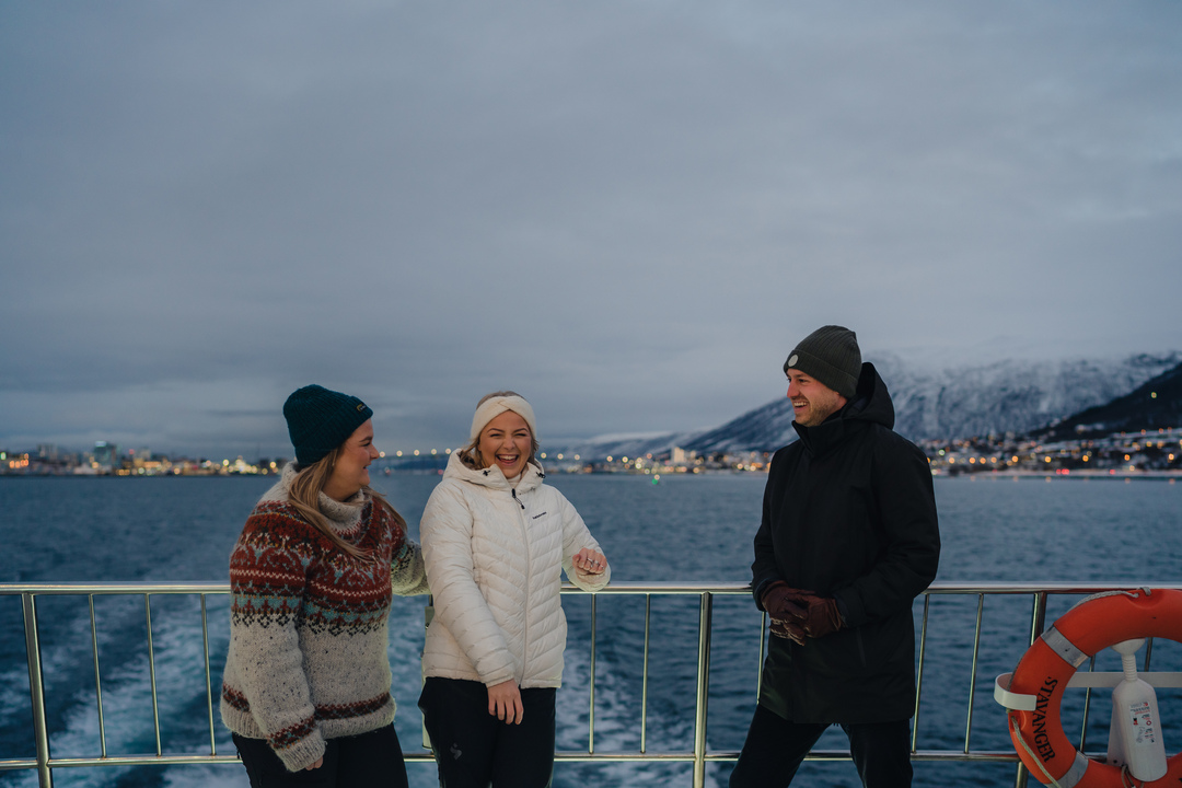 Group of smiling guests on the aft deck of a Rødne boat leaving Tromsø, with city lights, the bridge, and the Arctic Cathedral faintly visible in the background.